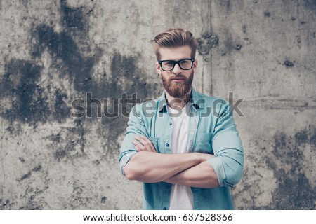 Nerdy handsome bearded young man is standing near the wall outdoors. He is wearing casual outfit and looks severe