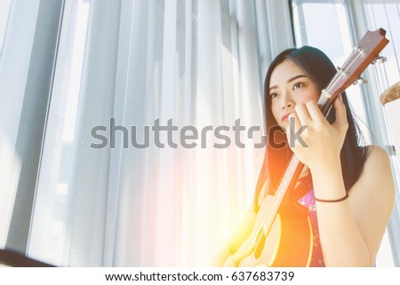 Happy young beautiful Asian woman musician enjoying music and playing ukulele indoors. Music and lifestyle concept