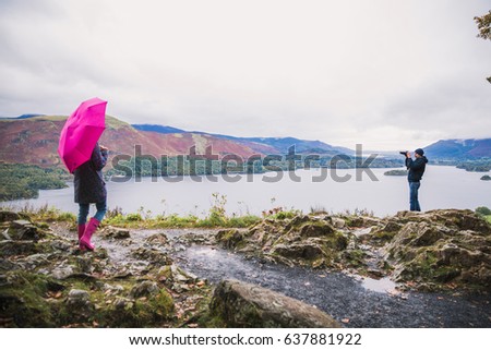 Young stylish blonde female stands at the top with pink umbrella and looks at amazing view. Young man stands at the top and does photo of amazing view on the photo camera