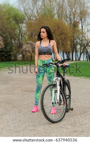 Girl beautiful brunette. Athletic body , pumped muscles. Bright colors , green ,pink . standing near a flowering tree . Sports bike , ride a bike along the shore