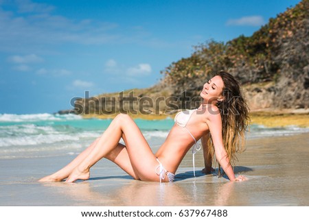 Beautiful young woman with long blond curly hair in white swimsuit sits on the sand of caribbean coastline at summer sunny day