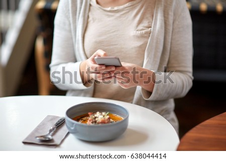 food, new nordic cuisine, technology, eating and people concept - woman sitting at cafe table with smartphone and bowl of vegetable pumpkin-ginger soup with goat cheese and tomato salad with yogurt