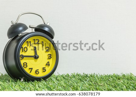 Closeup black and yellow alarm clock for decorate show a quarter to twelve o'clock or 11:45 a.m. on green artificial grass floor and cream wallpaper textured background with copy space