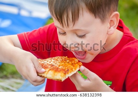 Cute little Caucasian kid eating pizza. Hungry child taking a bite from pizza on a pizza party. Smeared toddler. Pizza recipe.