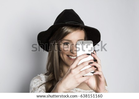 Smiling girl with black hat taking photo with phone, studio shot