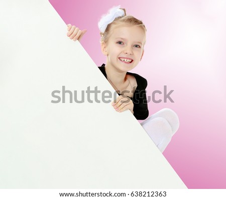 Beautiful little blonde girl dressed in a white short dress with black sleeves and a black belt.The girl peeks out from behind white banner.Close-up.Pale pink gradient background.