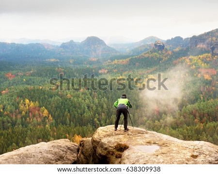 Hiker with camera on tripod takes picture from rocky summit. Alone photographer at edge photograph misty landscape, colorful forest in valley. 