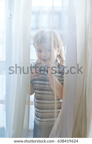 A little  boy dressed in a striped T-shirt is hiding behind a transparent curtain near the big window