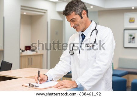 Happy mature doctor signing on form at reception in hospital. Handsome senior doctor filling patient form at medical clinic. Smiling doctor man reading patient case paper and making notes.