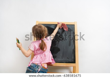 Little girl is cleaning blackboard or easel, she holding cleaning cloth in right hand and pencils in left hand