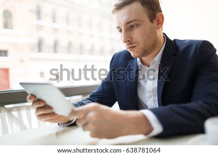 Portrait of modern businessman wearing formal suit using digital tablet at table in cafe and looking at screen