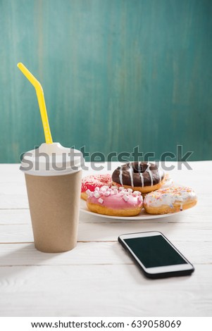 Unhealthy breakfast with coffee to go, plate of frosted donuts and smartphone wth black screen on wooden table. donuts and coffee background