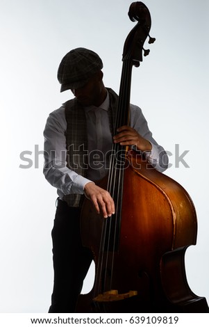 Silhouette of male musician playing contrabass. Wearing flat cap and plaid scarf, light grey background. Modern jazzman, blues, swing.
