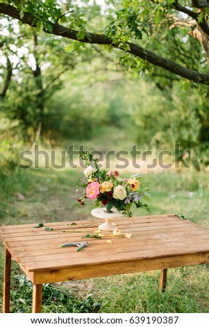 flower, summer and floral arrangement concept - desk florist on the summer glade among trees, beautiful fresh bouquet of white and yellow roses, peonies, red carnations, in white vase on wooden table