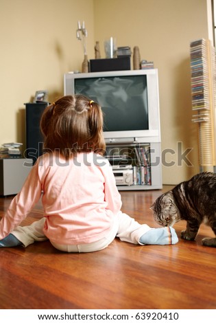 Rear view of little girl sitting on the floor and watching TV with cat near by