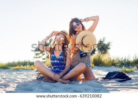 Two travel friends relaxing on  sunny beautiful beach, sitting on sand, making  pictures, talking and smiling.  Lifestyle and  holidays concept. 