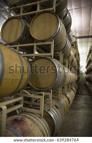 Vertical Close Up Front three-Quarter View, 4-High Row Stacked French Oak Wine Barrels, Cases of Wine in Background, Clean Commercial Lighted Warehouse, Cement Floor