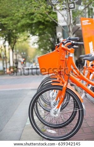 Blurred Backdrop for Copy Overlay -  Side Back to Front Horizontal View, Line Up Vibrant Orange Rental System Sharing Station Loaner Bikes Bicycles, Seats, Baskets, City Background - Portland, Oregon

