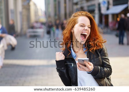 Exuberant young woman cheering at good news on her mobile phone and punching the air with her fist on an urban street with copy space