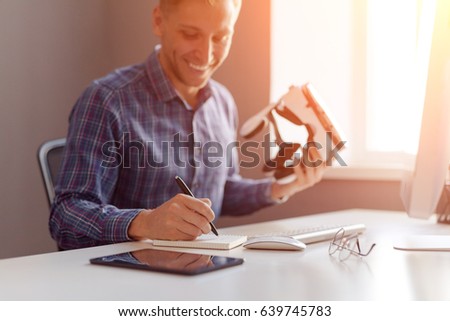 Smiling office worker writing in notepad and holding the VR goggles. Horizontal indoors shot. 