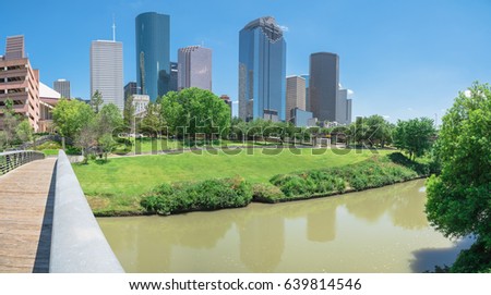 Panorama view downtown Houston taken from pedestrian bridge at daytime, cloud blue sky. Green lawn, trees, modern skylines along Bayou river. Most populous city in Texas, fourth-most in United States.