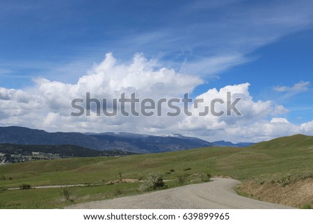 Gravel road through green grass field and hills with mountains in background.