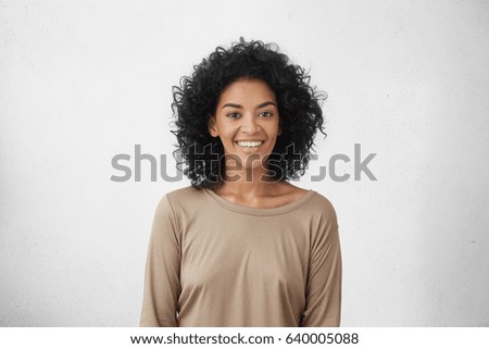 Waist up portrait of cheerful young mixed race female with curly hair posing in studio with happy smile. Dark-skinned woman dressed casually smiling joyfully, showing her white straight teeth Royalty-Free Stock Photo #640005088