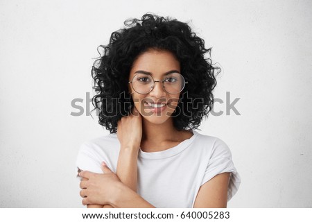 Headshot of attractive young mixed race woman wearing big spectacles and white top holding hand behind her neck and looking at camera with subtle smile during awkward conversation with some guy