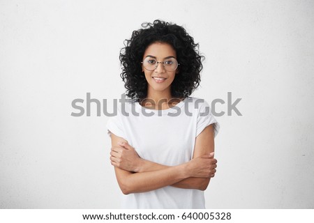 Body language. Isolated studio portrait of constrained young dark-skinned woman in round eyewear standing at white wall with arms folded, looking with subtle smile while visiting her therapist