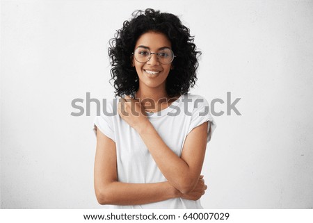 Attractive mixed race female actress wearing round eyeglasses and t-shirt looking and smiling at camera in closed posture, keepings arm on her breast, standing against white wall during audition