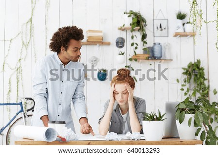 Team of young designers facing deadline while working on architectural project at office. Senior African american engineer checking work of sad trainee girl, pointing at her mistakes in blueprint