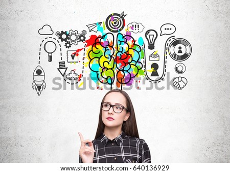 Close up of a nerdy girl wearing a checkered shirt and glasses. She is standing near a concrete wall with a start up sketch and a colorful brain icon