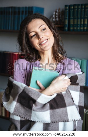 Portrait of a happy mature woman covered in blanket holding book and looking away indoors