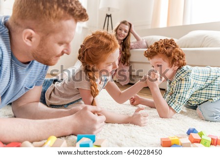 side view of brother and sister playing arm wrestling with parents near by, family fun at home concept