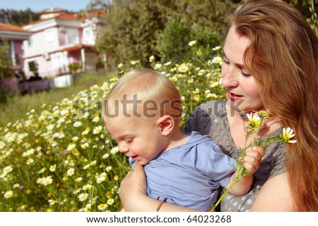 young looking grandmother with red hair holding her grandson in the field of daisies on the sunny summer day.