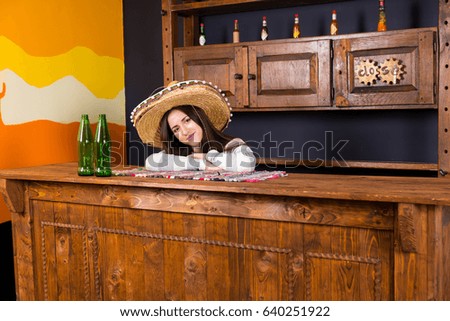 Beautiful young woman in a sombrero leaned on bar counter with beer bottles in Mexican pub