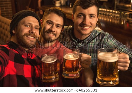 Three handsome young men taking a selfie at the bar holding beer glasses. Male friends enjoying drinking beer together at the local pub taking pictures bachelor party event masculinity mood festive