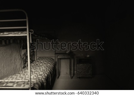Black and white picture of dark empty prison cell with iron bunk bed and bedside table with aluminum dishes