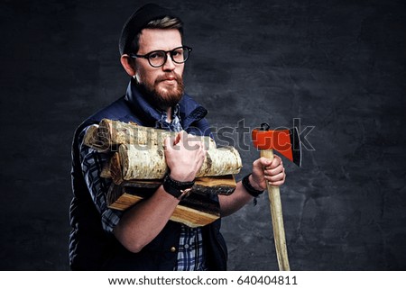 Alternative bearded male in eyeglasses dressed in fleece shirt holds firewoods and axe over grey background.
