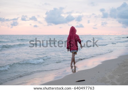 A boy wearing a red shirt is walking by the sea in the evening