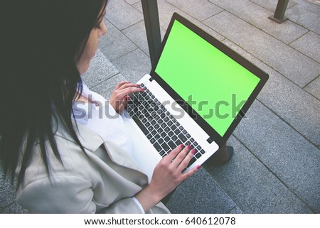 Useful gadget for business. Low angle closeup view of beautiful business woman in smart casual wear working on laptop while sitting outdoors. Green chroma key screen on laptop.