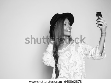 Beautiful girl with long shiny hair and hat taking selfie with the phone, studio shot, black and white photo