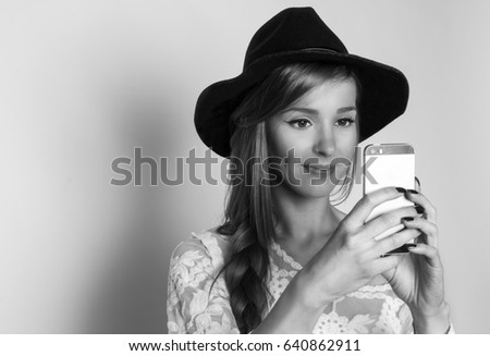 Beautiful girl with hat taking picture with smart phone, studio shot, black and white photo