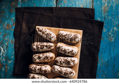 Cakes eclairs in white chocolate and chocolate crumbs on a wooden board on a brown napkin on a worn old blue table. View from above