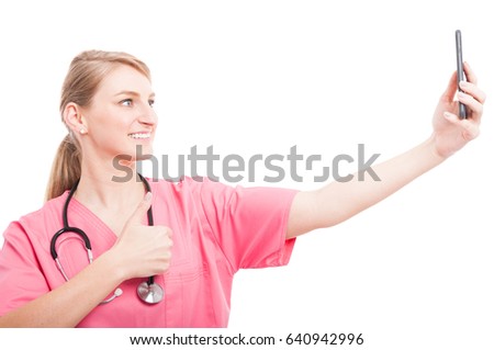 Nurse wearing scrubs taking selfie with smartphone showing like and smiling isolated on white background