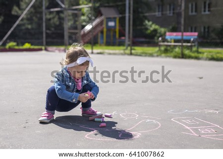 Little beautiful girl in a jeans jacket draws with colored chalks on the playground