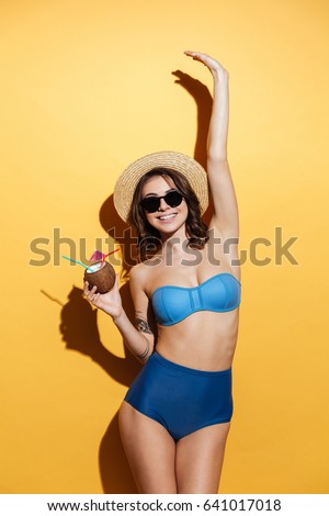Image of cheerful young woman in swimwear isolated over yellow background holding cocktail. Looking at camera.