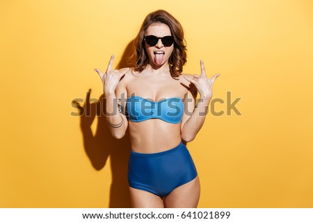 Image of screaming young woman in swimwear isolated over yellow background. Looking at camera make rock gesture.
