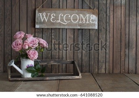 Wooden sign Welcome and Beautiful pink roses in a vintage jug on rustic wooden background