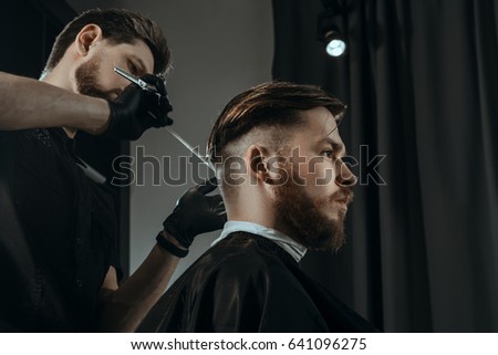BARBERSHOP THEME. BEARDED BARBER IN BLACK RUBBER GLOVES IS TRIMMING THE HAIRCUT OF HIS YOUNG SERIOUS CLIENT. HE IS USING A HAIR CLIPPER Royalty-Free Stock Photo #641096275